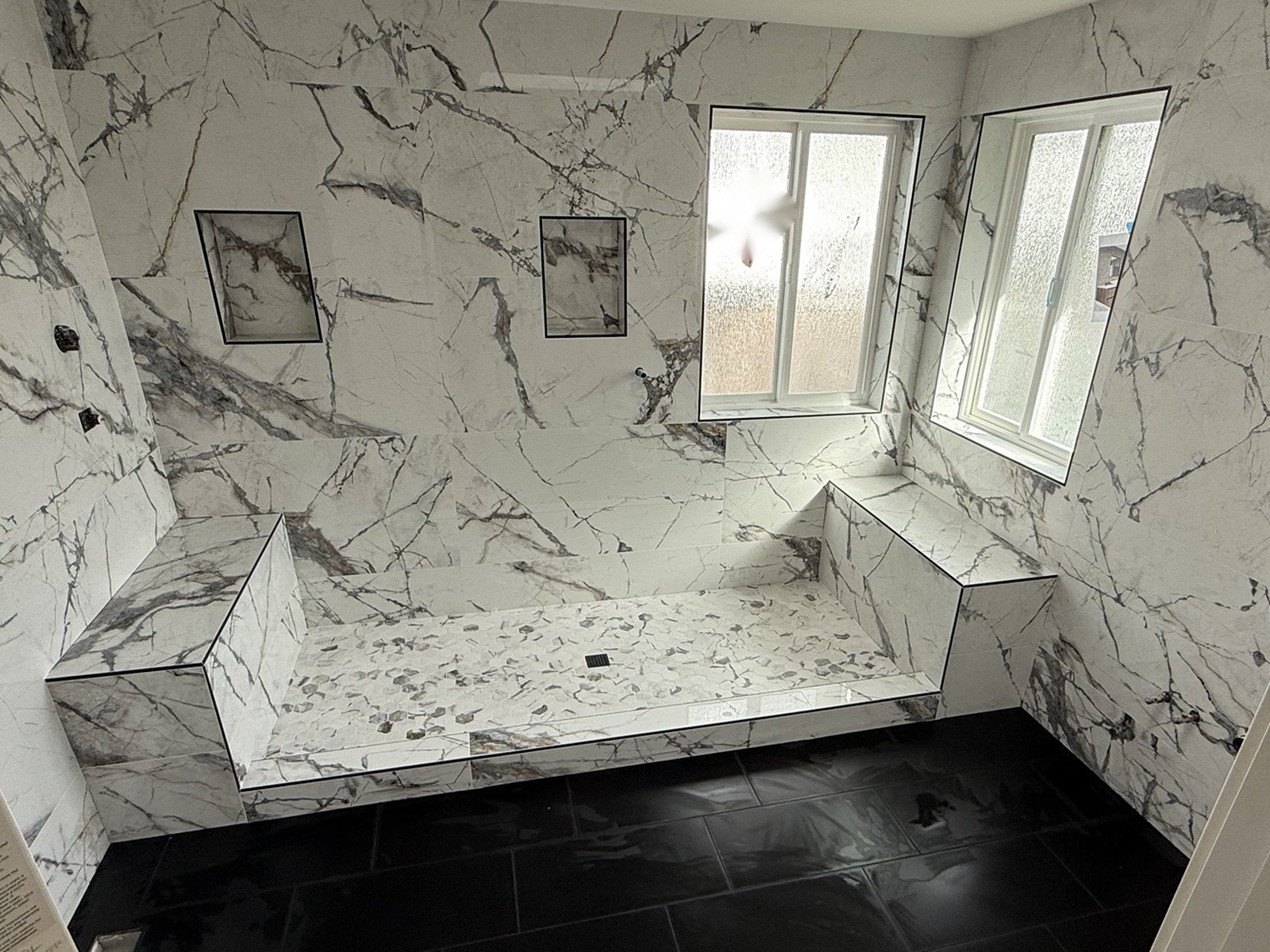 After: walk-in shower with large format marble-look tile, two tiled benches, twin niches and a black tile floor